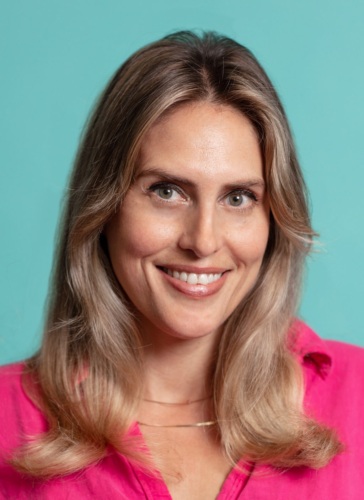 A smiling woman with long blonde hair wearing a bright pink shirt against a teal background.